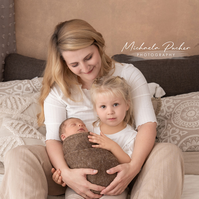 Mama sitzt auf dem Sofa und hält gemeinsam mit ihrer kleinen Tochter das schlafende Neugeborene auf dem Schoß; beide Kinder sind in natürliche Töne gekleidet.